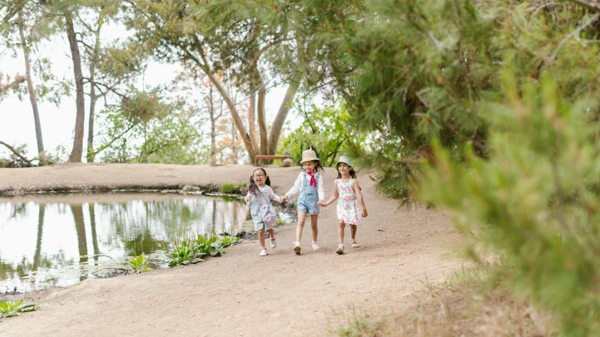 Three children holding hands and laughing while walking by a serene pond in a lush green park.