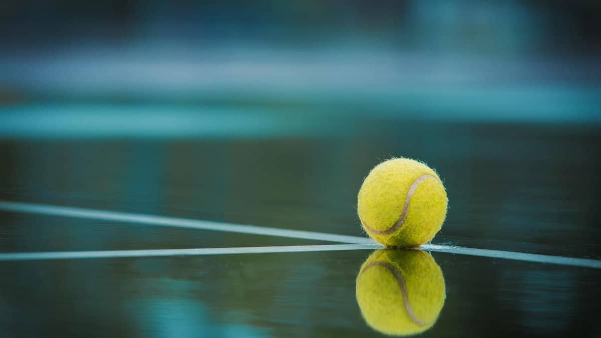 A tennis ball sits on a wet court with its reflection visible, capturing a serene sports moment.