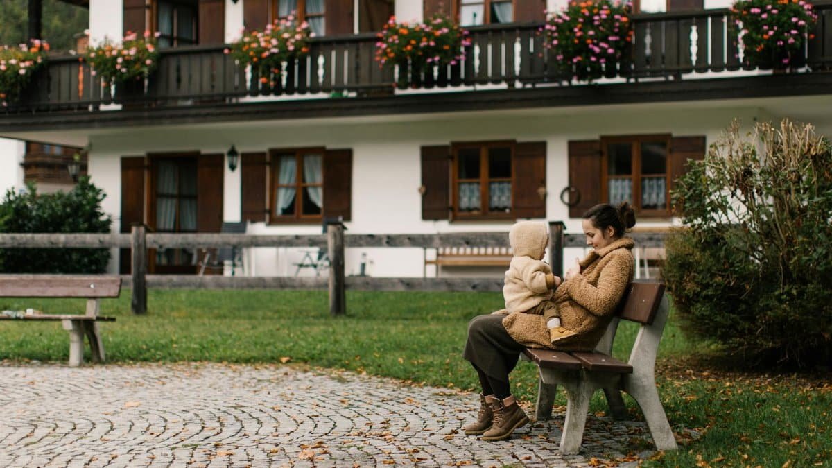 A mother and child in warm coats enjoy a moment on a bench outside a charming home with flowered balconies.