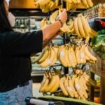 A woman in a face mask selects fresh bananas in a supermarket produce section, emphasizing safe shopping practices.