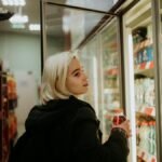 Blonde woman selecting a beverage from a refrigerated shelf at a convenience store.