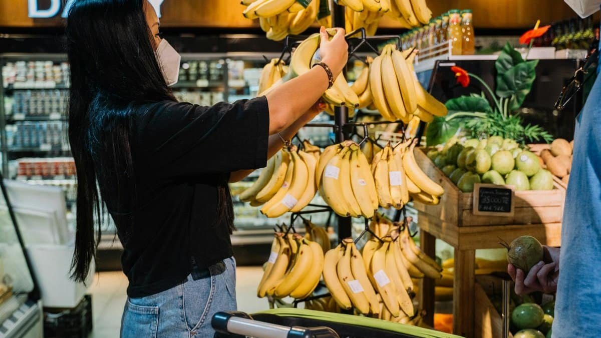 A woman in a face mask selects fresh bananas in a supermarket produce section, emphasizing safe shopping practices.