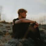 A young man seated on frosty grass, enjoying a serene sunset outdoors.