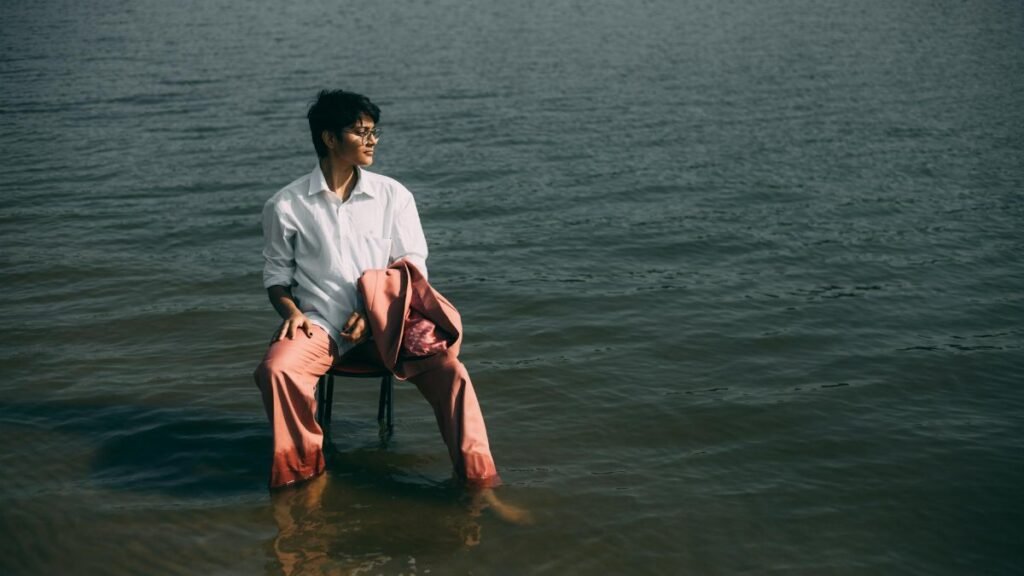 A man wearing a white shirt and pink suit pants sits in the water near a lake, exuding a sense of solitude and style.