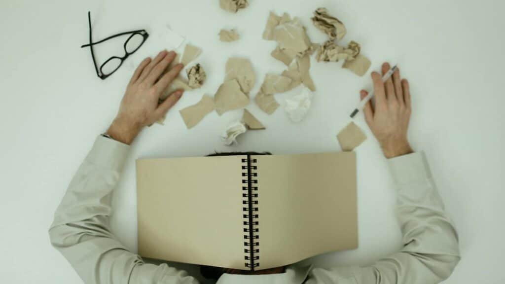 Overhead view of an exhausted man resting with a notepad over his head and crumpled papers around.