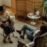 Two women engaged in a psychotherapy session in a warm, inviting interior with plants and natural lighting.