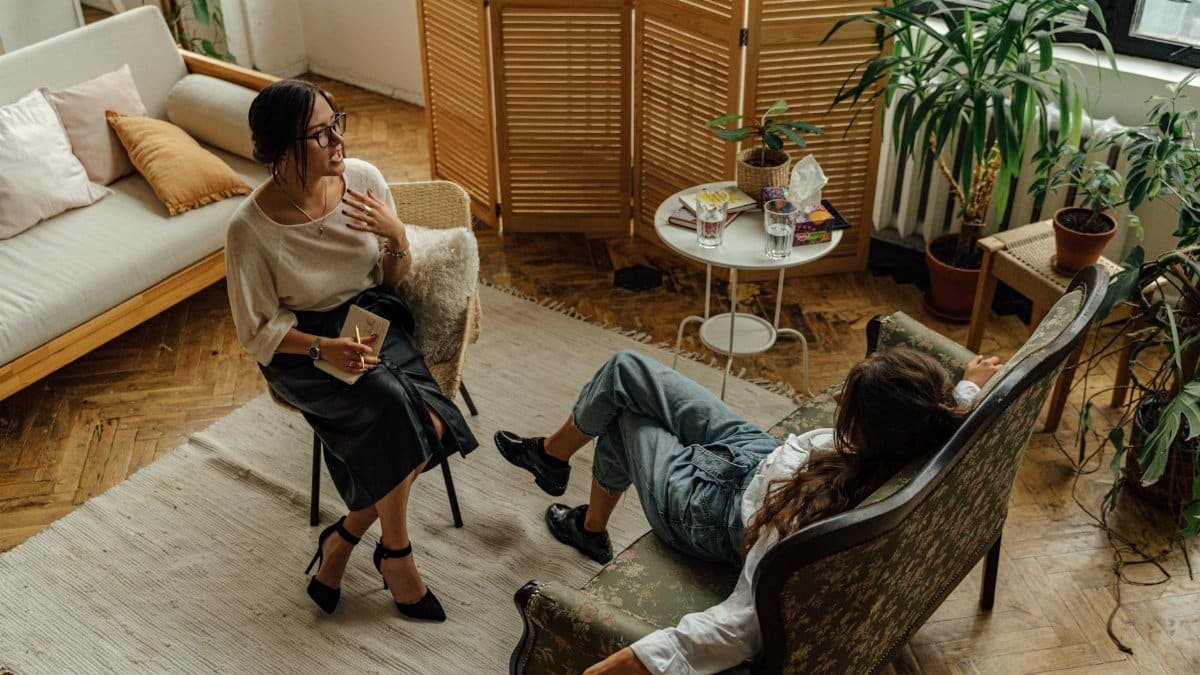 Two women engaged in a psychotherapy session in a warm, inviting interior with plants and natural lighting.
