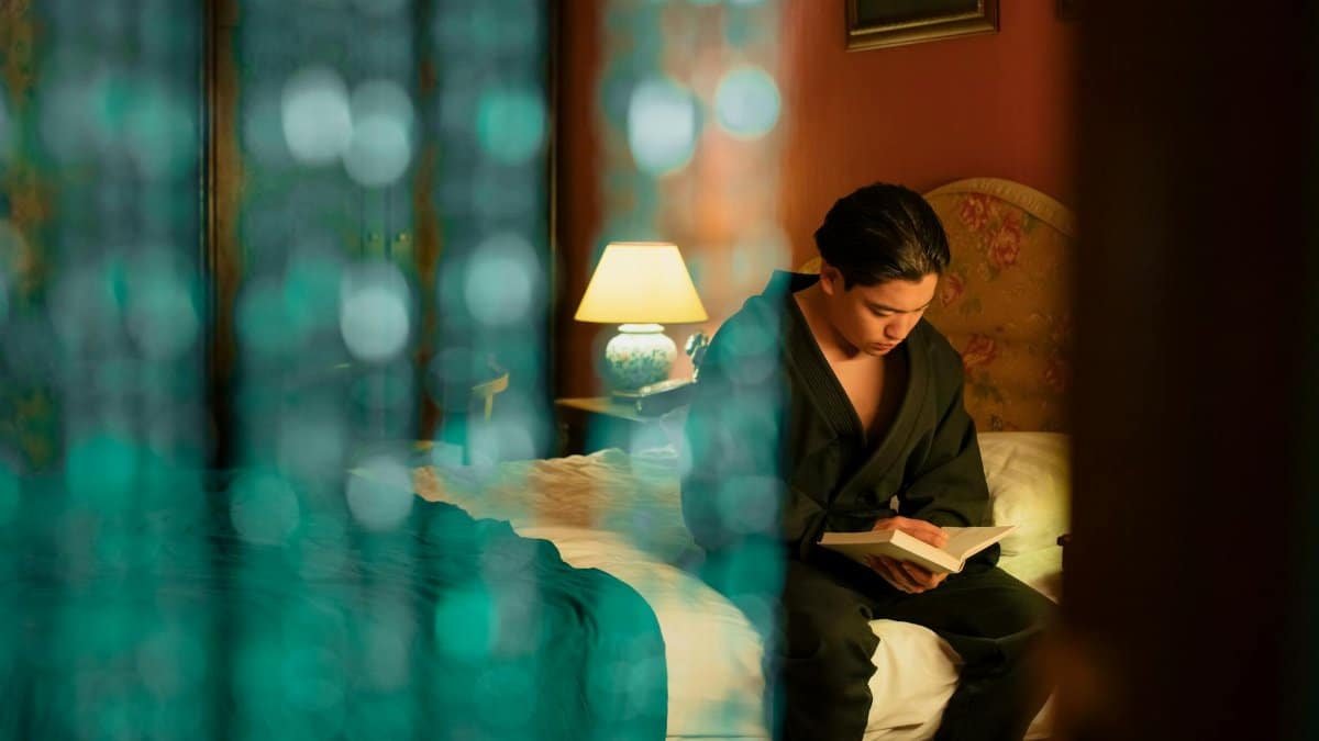 Young man in a robe reading a book in a warmly lit hotel room, blurred beads in the foreground.