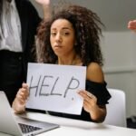 An unhappy businesswoman showing a sign reading 'HELP' in an office setting.