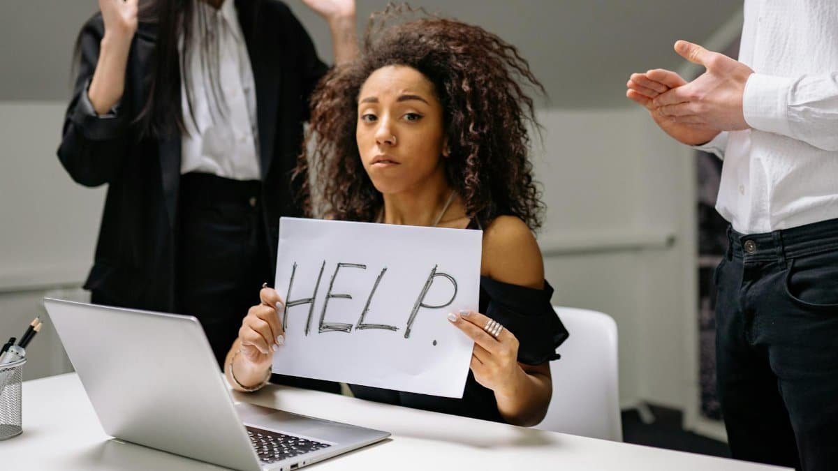 An unhappy businesswoman showing a sign reading 'HELP' in an office setting.