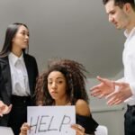 A woman in an office holds a 'HELP' sign while facing conflict with coworkers.