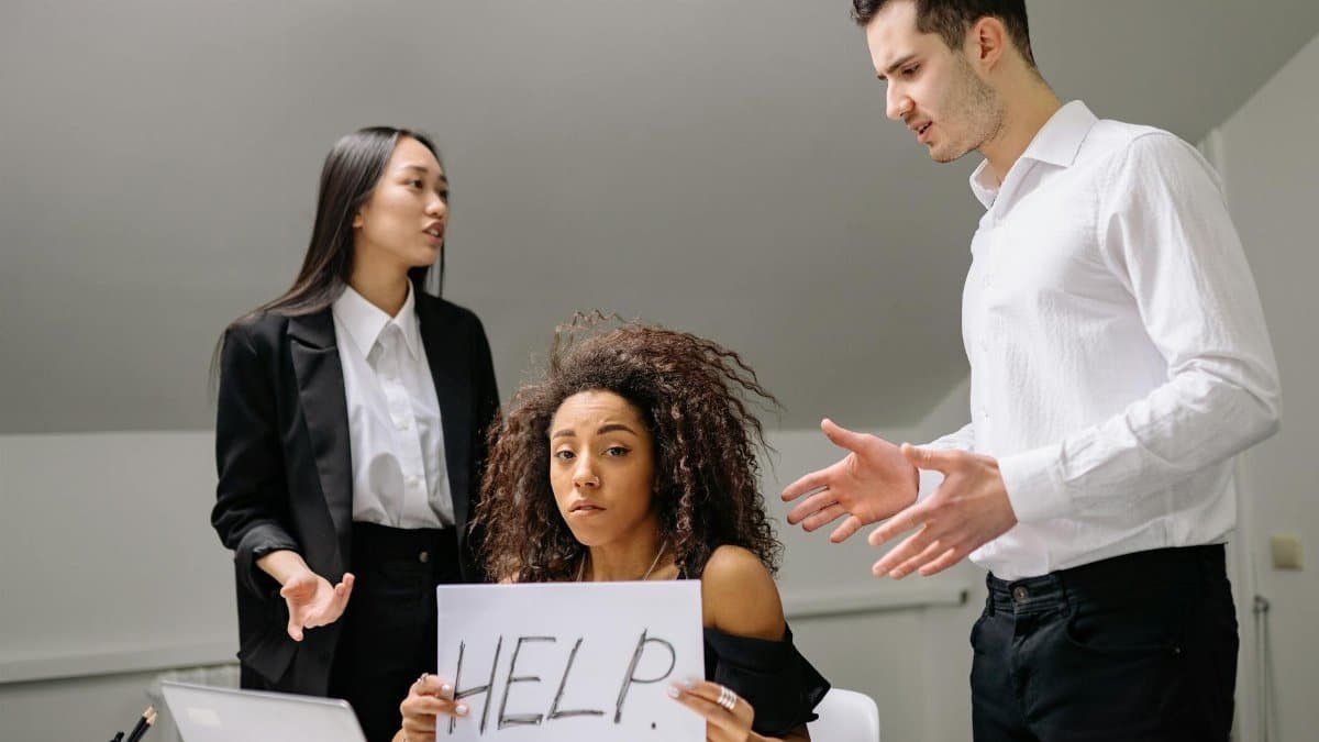 A woman in an office holds a 'HELP' sign while facing conflict with coworkers.