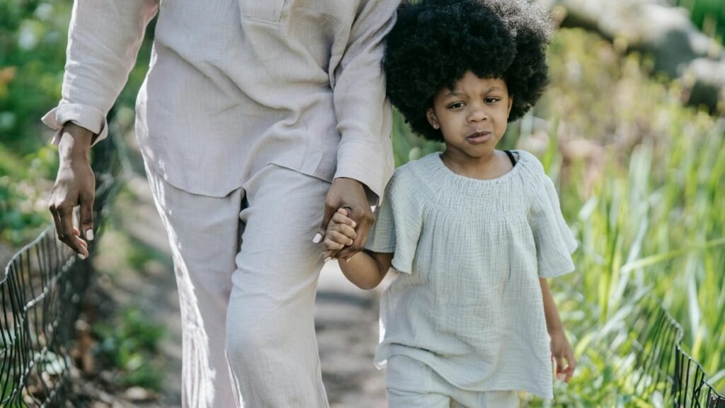 A young girl with curly hair holds hands with an adult, walking along a garden path.
