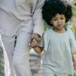 A young girl with curly hair holds hands with an adult, walking along a garden path.