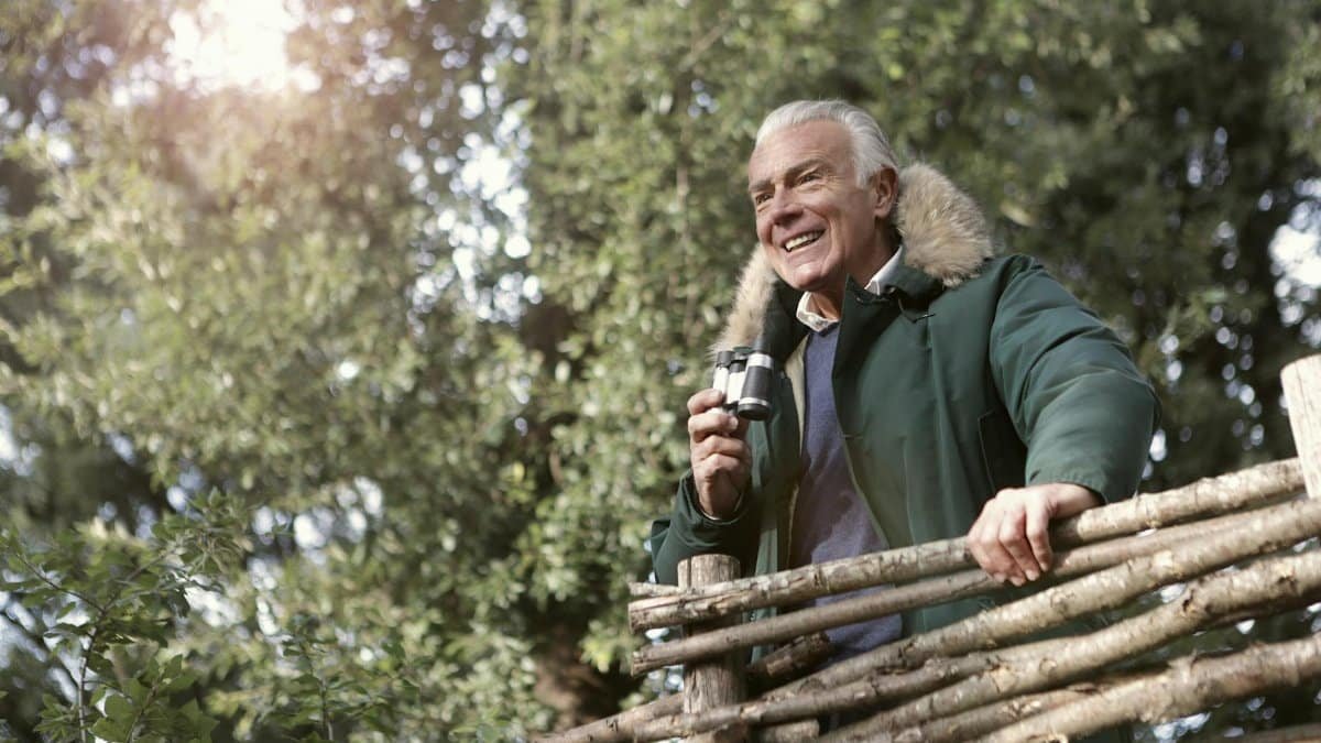 Elderly man enjoying a sunny day birdwatching in a park with binoculars.