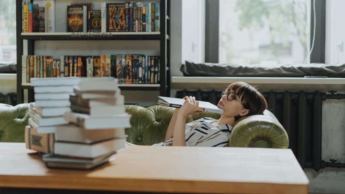 A woman lounging on a sofa with a book, surrounded by stacks of books in a cozy library.
