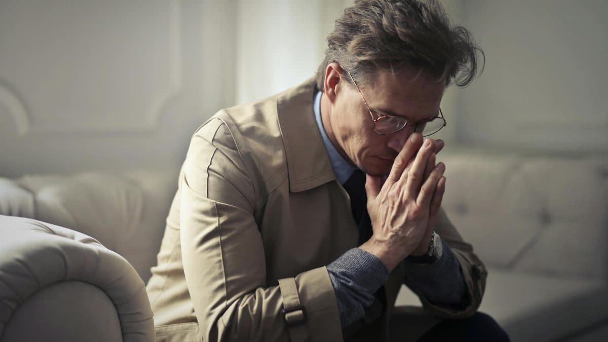 Side view of upset male entrepreneur in formal wear sitting on couch in modern living room and contemplating about financial problems while leaning on hands and looking down