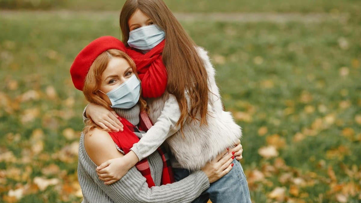 A mother and daughter wearing masks embrace in a fall park, showcasing warmth and protection.