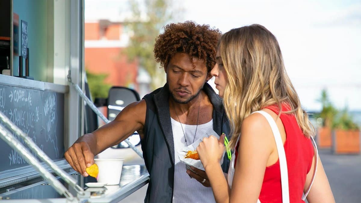Hungry young black man and woman in casual clothes eating yummy crisps with sauce and hamburger while having lunch in food truck