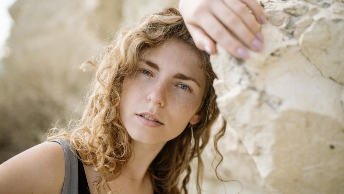 A contemplative young woman with curly hair leaning against a rock in a natural setting.