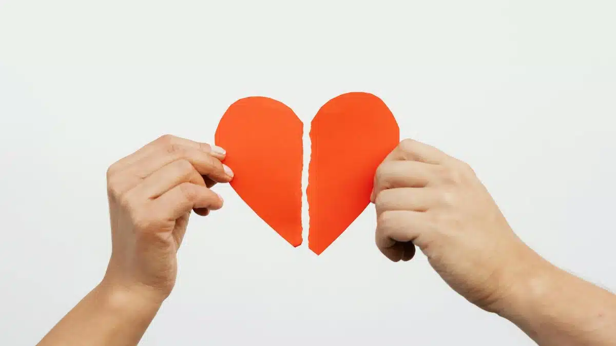 A close-up view of hands holding a torn red paper heart, symbolizing heartbreak or relationship issues.