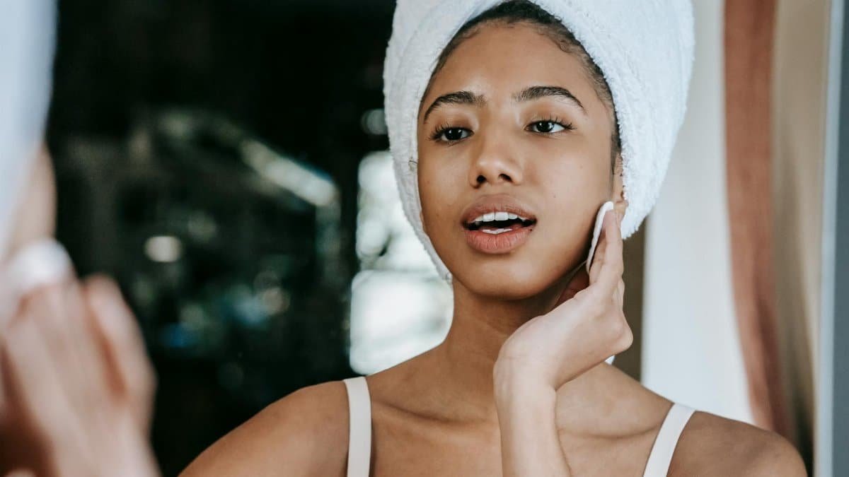Ethnic female with mouth opened cleaning face with cotton pad while looking at mirror during daily beauty care