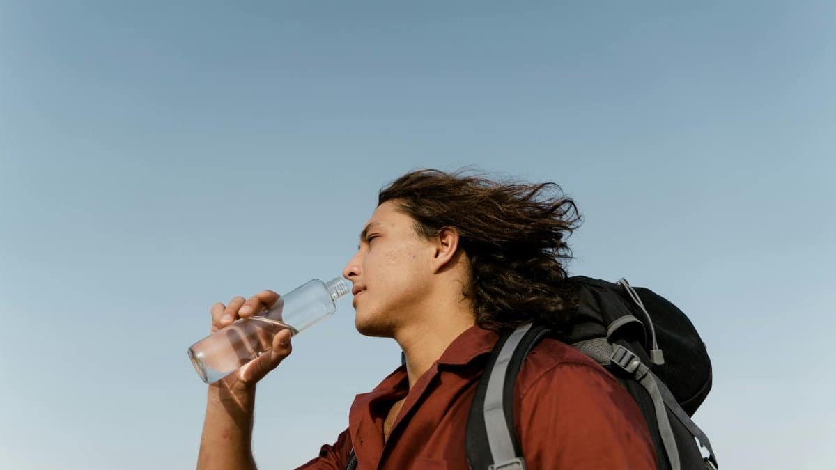A young man drinking water while hiking with a backpack under the clear sky.