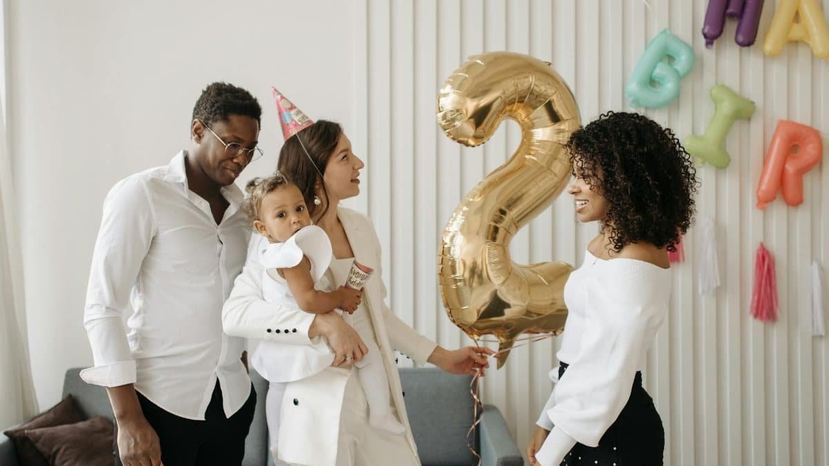 A joyful family gathers with balloons and party hats for a child's second birthday celebration indoors.