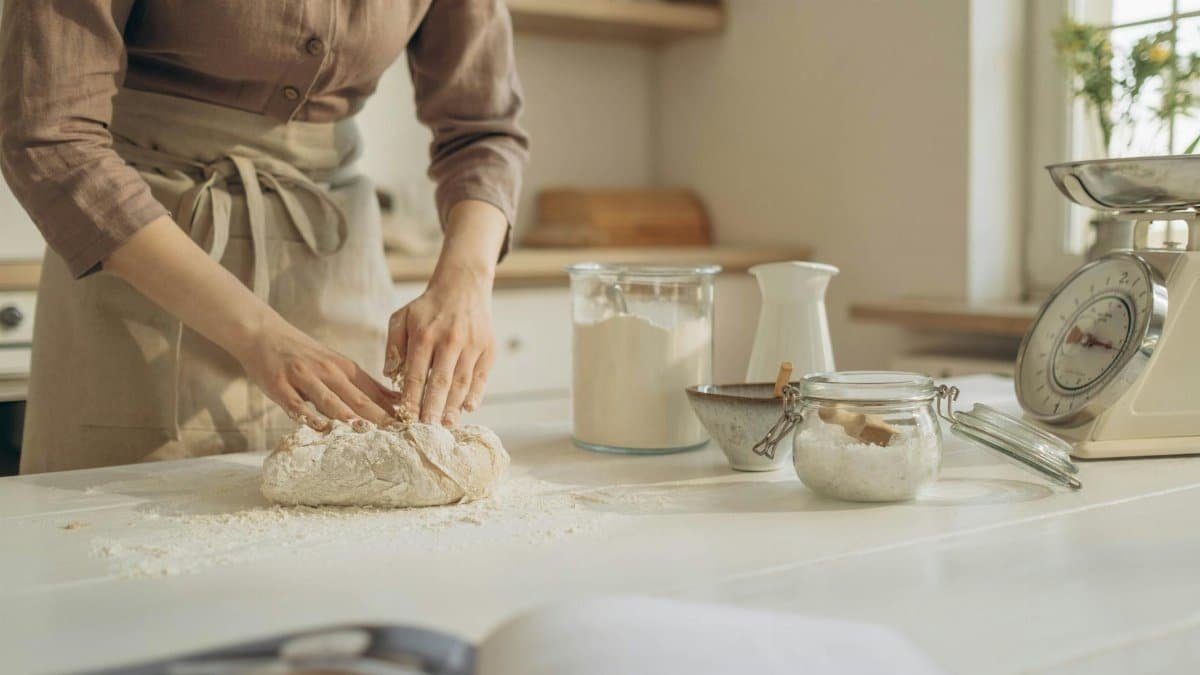 A person is kneading dough on a white kitchen countertop, surrounded by baking ingredients and tools.