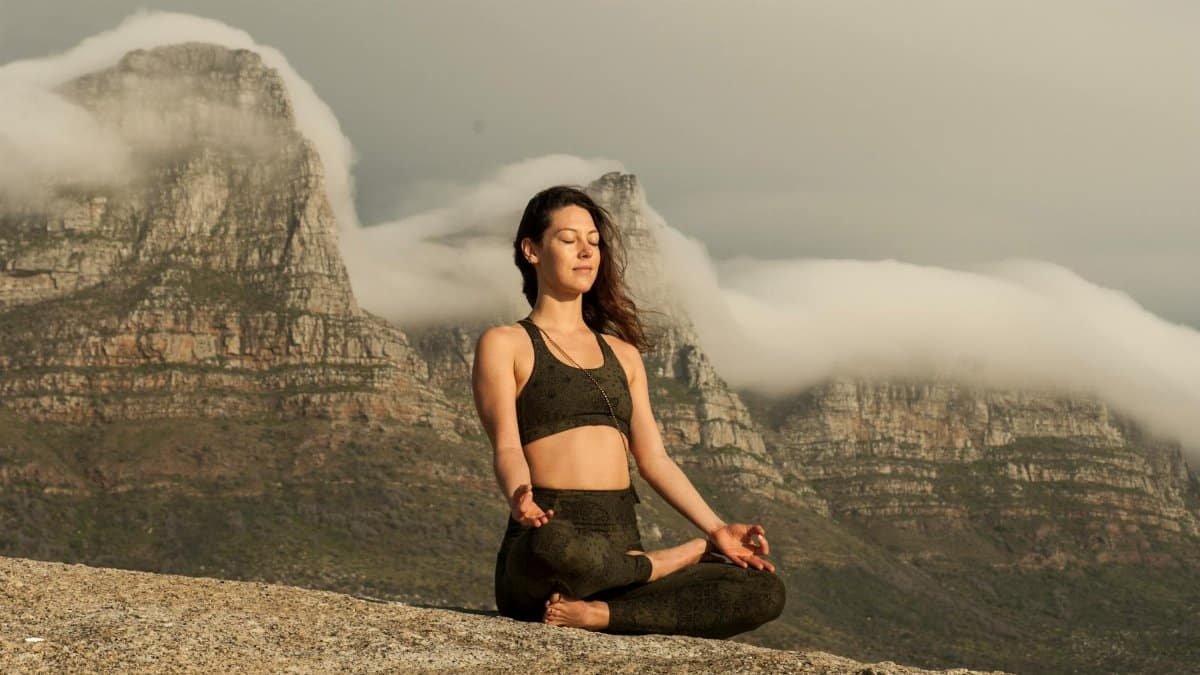 A woman practicing yoga and meditation outdoors in a scenic mountain setting.