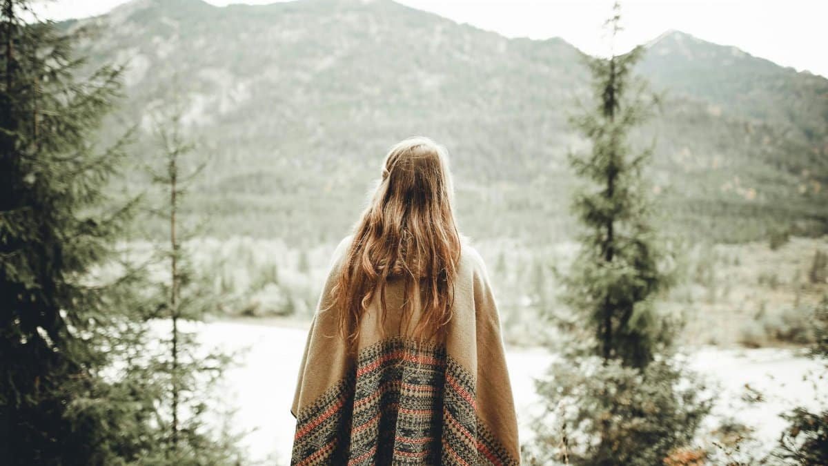 A woman with long hair wrapped in a blanket admires the scenic winter forest and mountain view.