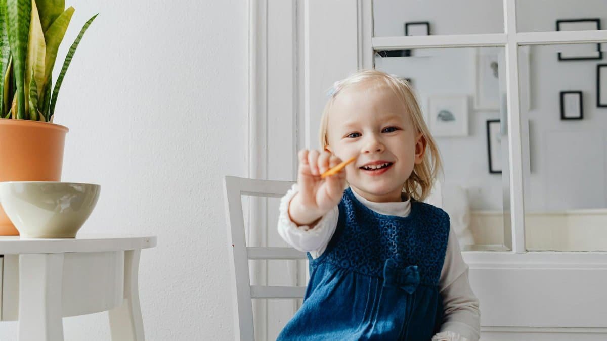 A cheerful young girl smiling while holding out a biscuit indoors.