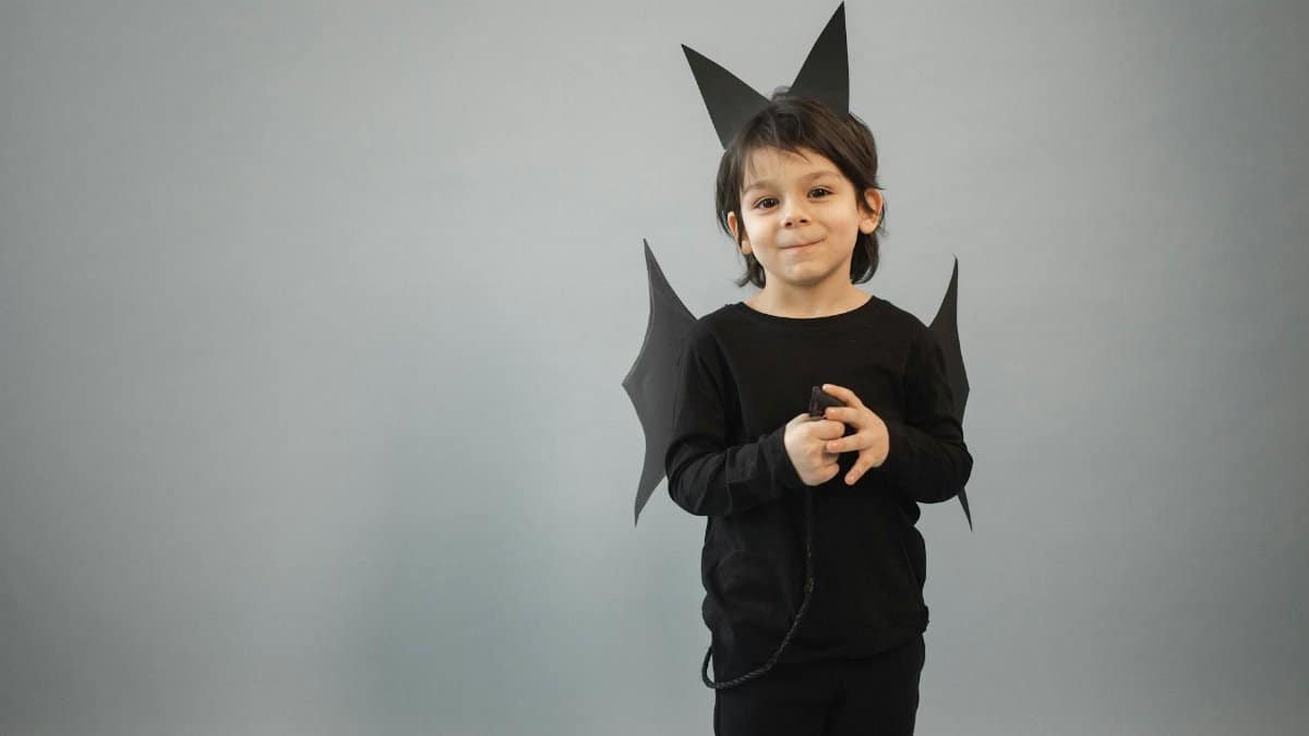 Positive boy in black bat outfit with wings and ears looking at camera on gray background with tail in hands