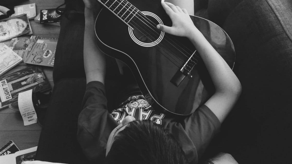 A young boy lying indoors on a couch playing an acoustic guitar with scattered books around.