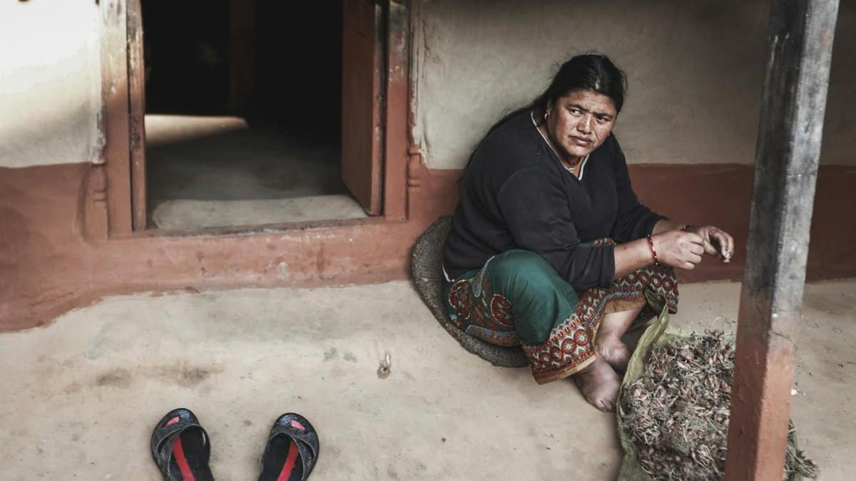 An adult woman sits by a rustic doorway sorting dried crops in a traditional setting.
