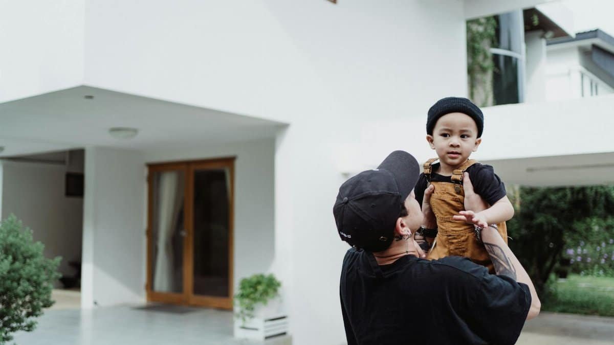 A father playfully lifting his child outside a modern home on a sunny day.