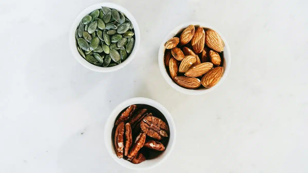 A top-down view of assorted nuts in bowls, featuring almonds, pumpkin seeds, and pecans, on a marble surface.