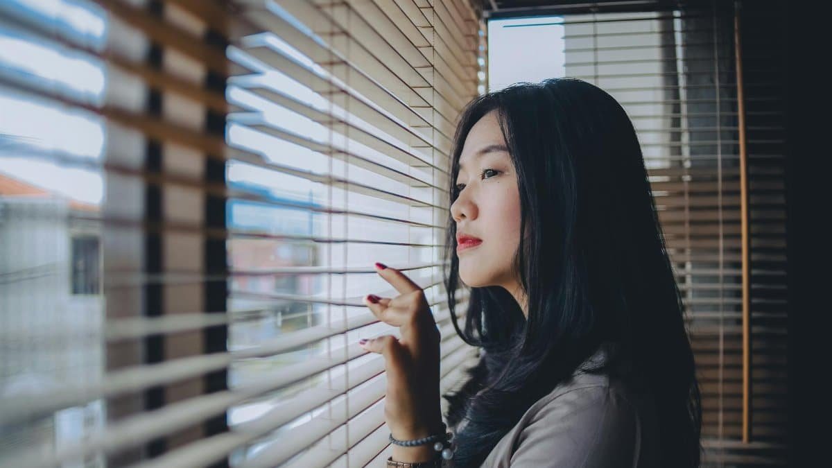 A thoughtful Indonesian woman looks out through window blinds, contemplating indoors.