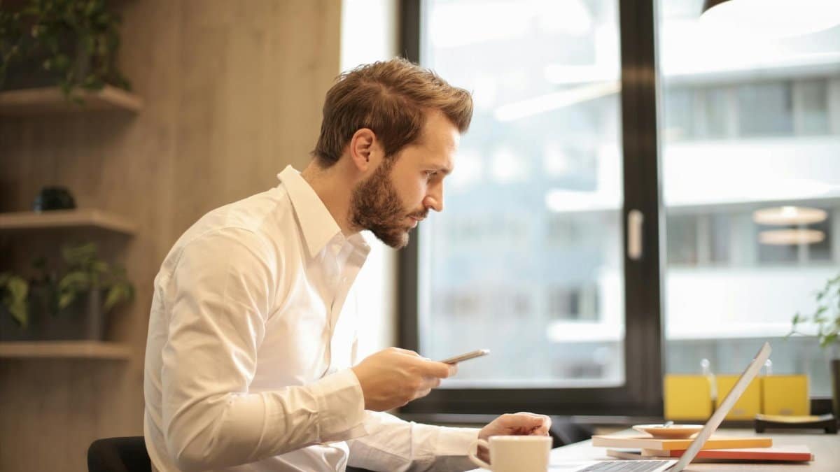 Focused businessman working on laptop while checking smartphone in modern office.