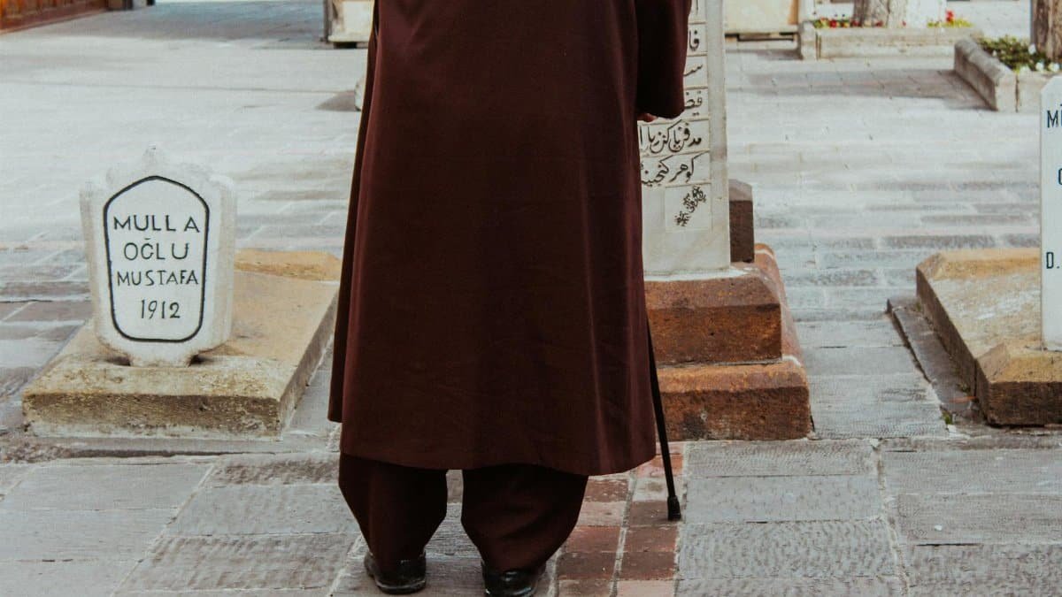 An elderly man in traditional clothing stands at a historical cemetery reading gravestones.