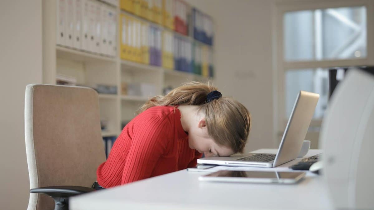 A tired woman in a red sweater leans her head on a desk with a laptop, symbolizing workplace fatigue.