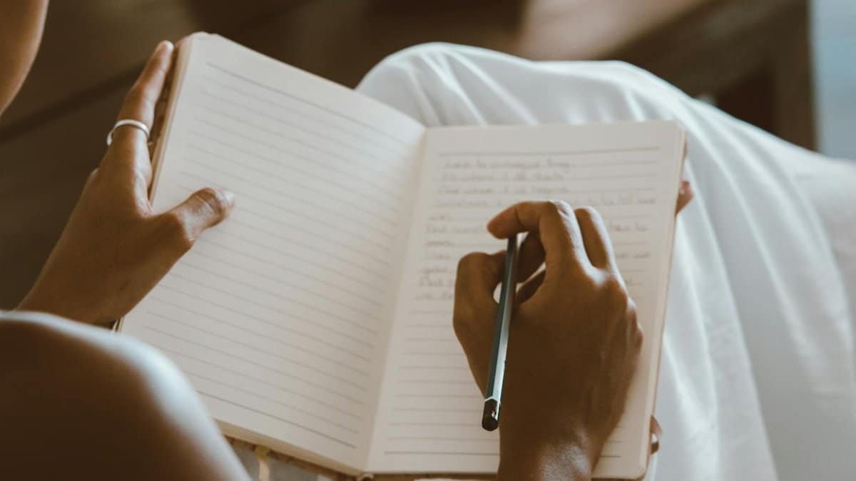 Close-up of hands writing in a journal with a pencil on a seated lap.