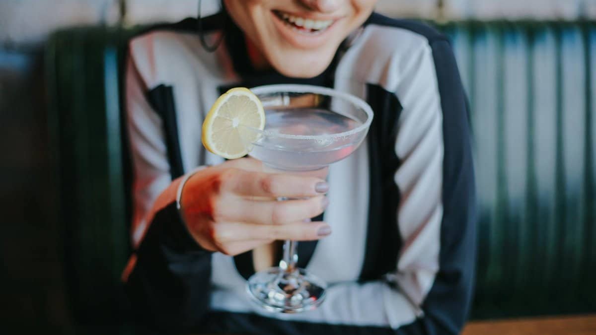 A woman smiles while holding a lemon-garnished cocktail at a cozy restaurant.