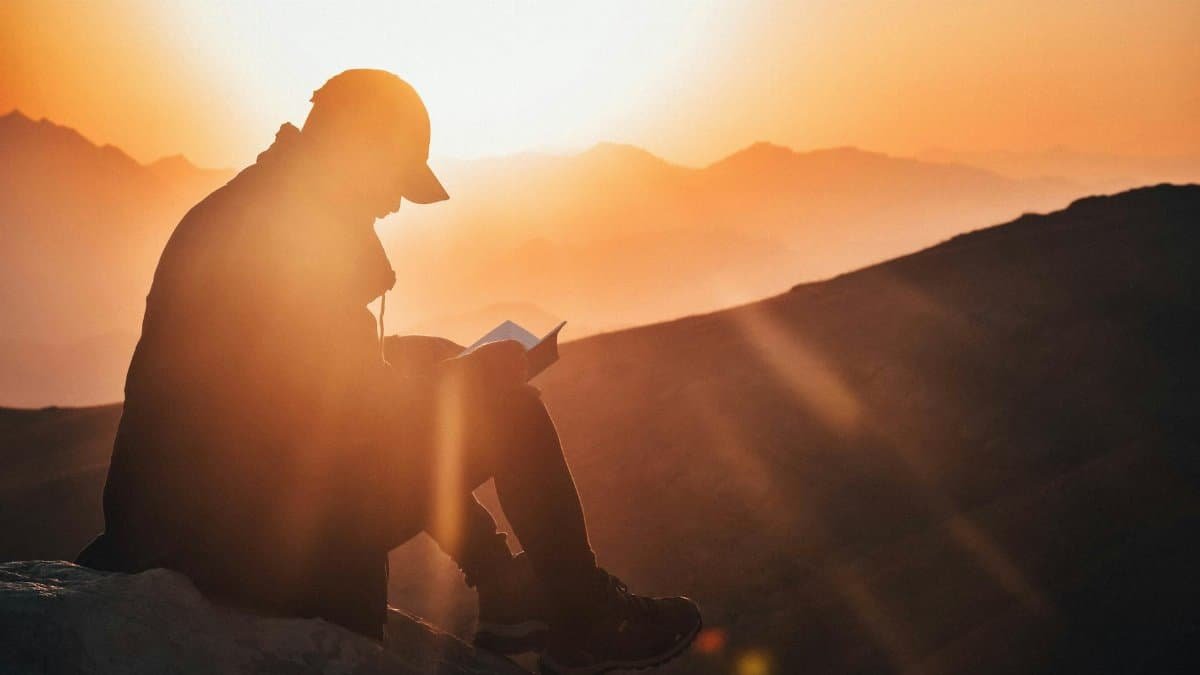 A man reads a book silhouetted against a vibrant sunset, seated on a mountain edge.