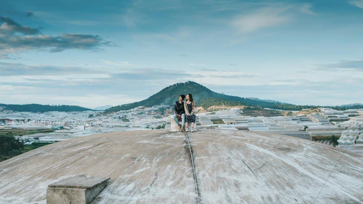 A couple sits atop a large concrete structure, overlooking a scenic mountain landscape under a blue sky.