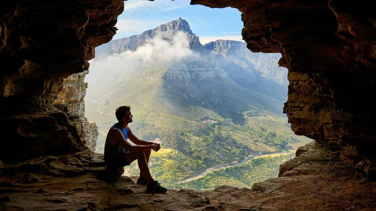 A man sitting in a cave overlooking a majestic mountain landscape under daylight.