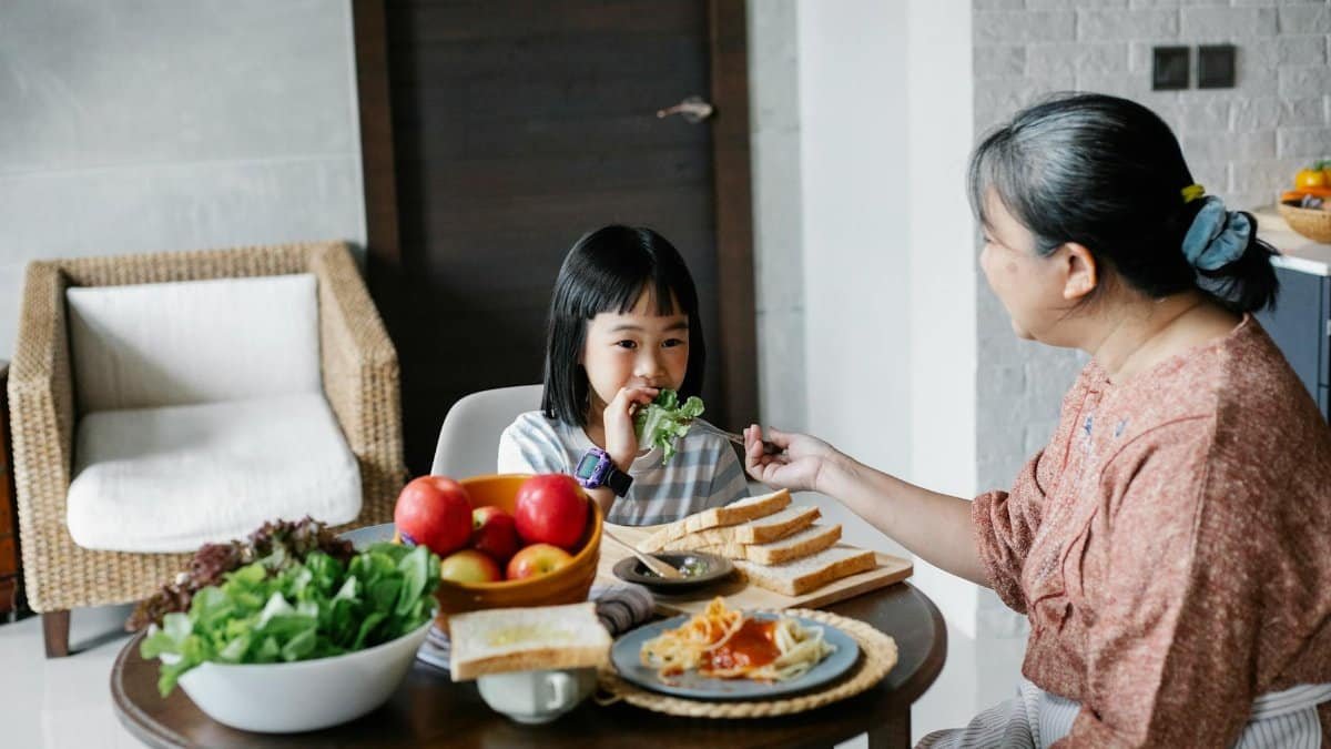 Ethnic daughter eating lettuce leaf while having healthy meal with mother in modern apartment