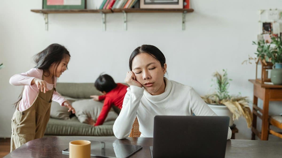 A tired mother working remotely on her laptop while children play in the background.