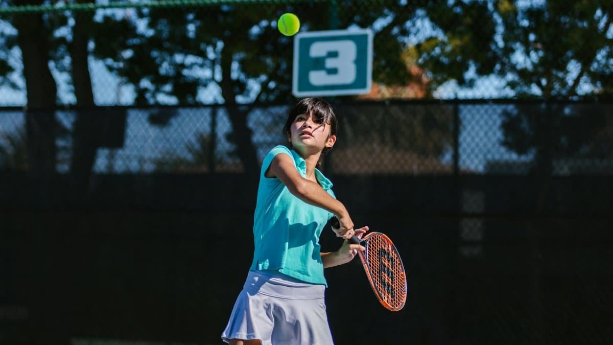 Teenage girl returns tennis ball with backhand swing on outdoor court.