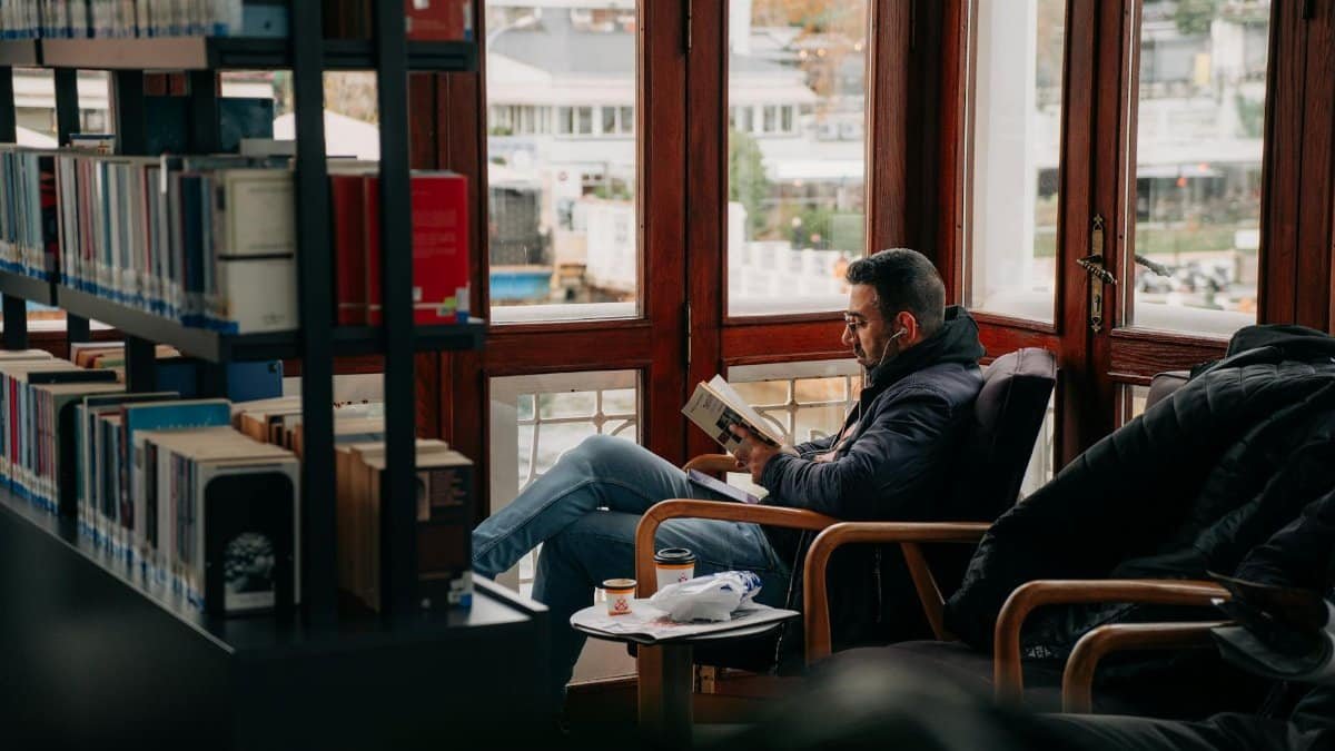 Adult man reading a book in a cozy library with large windows and shelves.